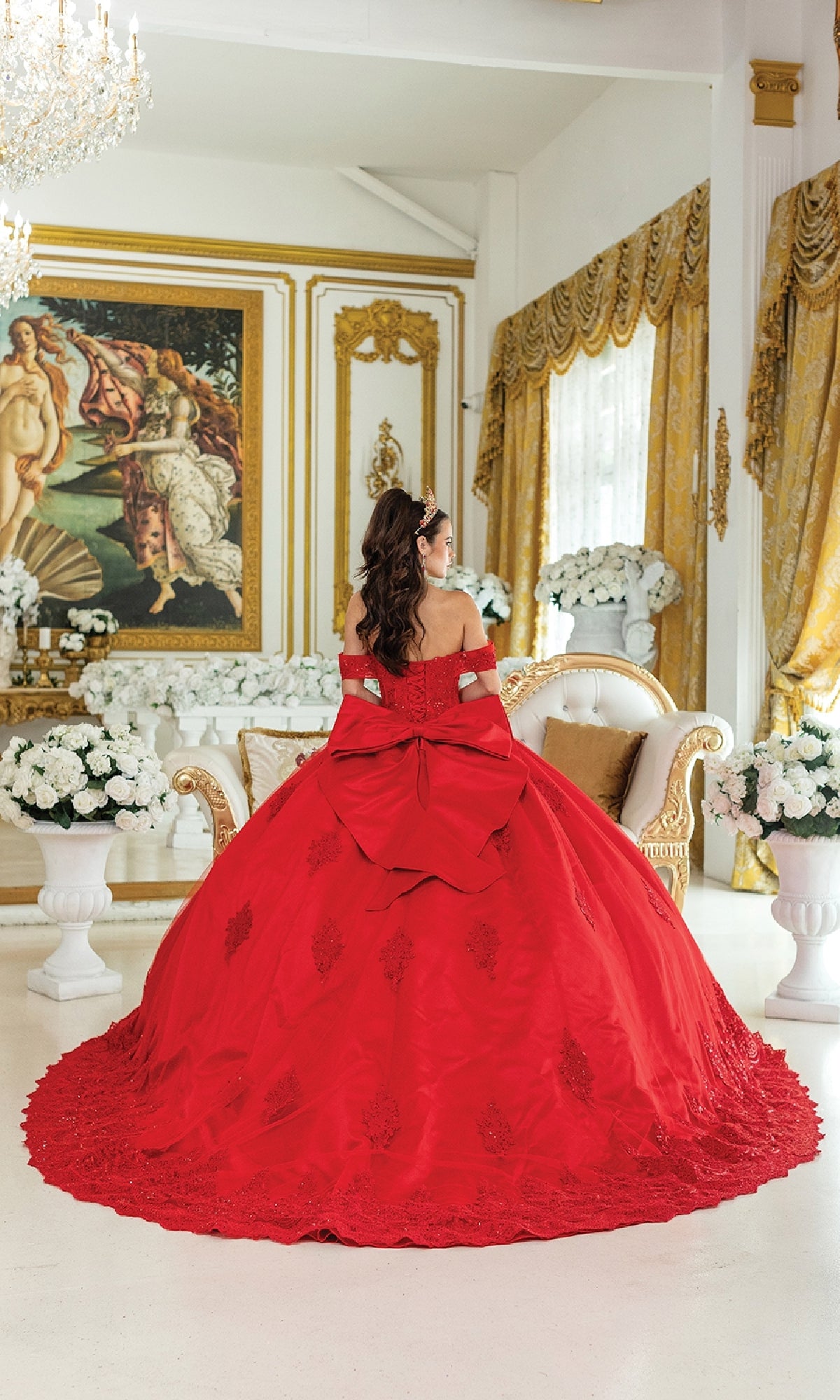 A woman in the Quinceanera Dress 1913 by Dancing Queen, an off-the-shoulder red ball gown with a corset bodice, sits on an ornate sofa in a lavish room adorned with gold accents, white flowers, and a classical painting.